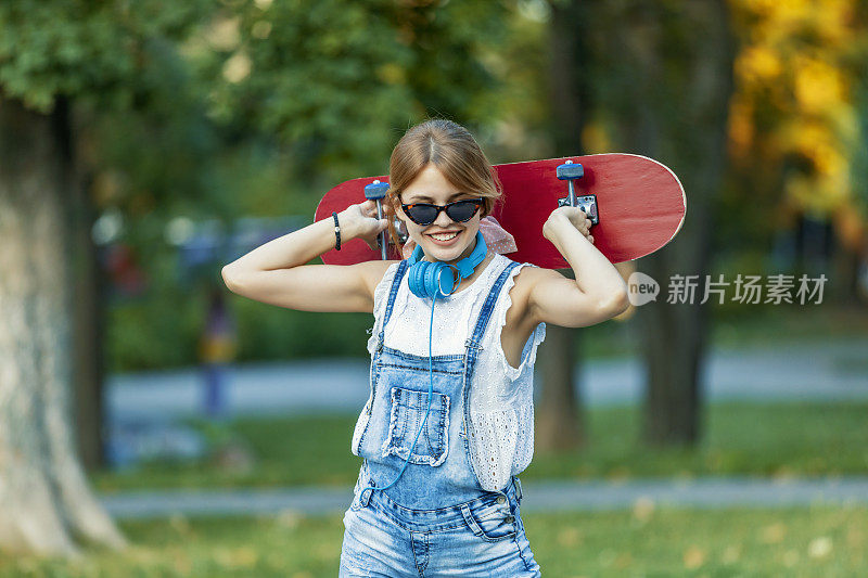 在一个阳光明媚的日子里，在公园里玩滑板的漂亮年轻女子。年轻迷人的美丽和时尚的年轻女人与滑板摆姿势的照片。春天的一天，她站在城市的街道上享受。快乐休闲的着装