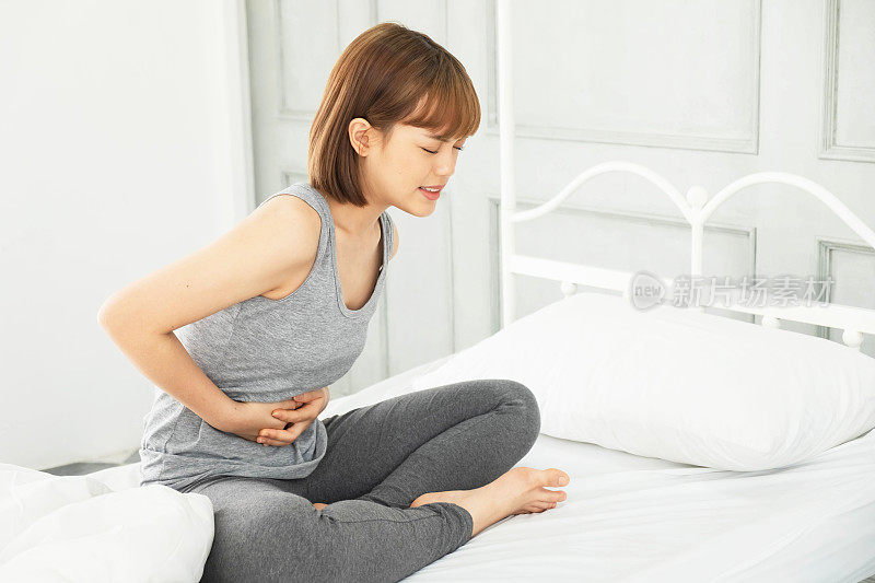 年轻美丽的亚洲女人穿着灰色的衣服，早上醒来在白色的床上，身体疼痛，拥抱的手在腹部蜷缩，腹痛，腹痛，痛，痛