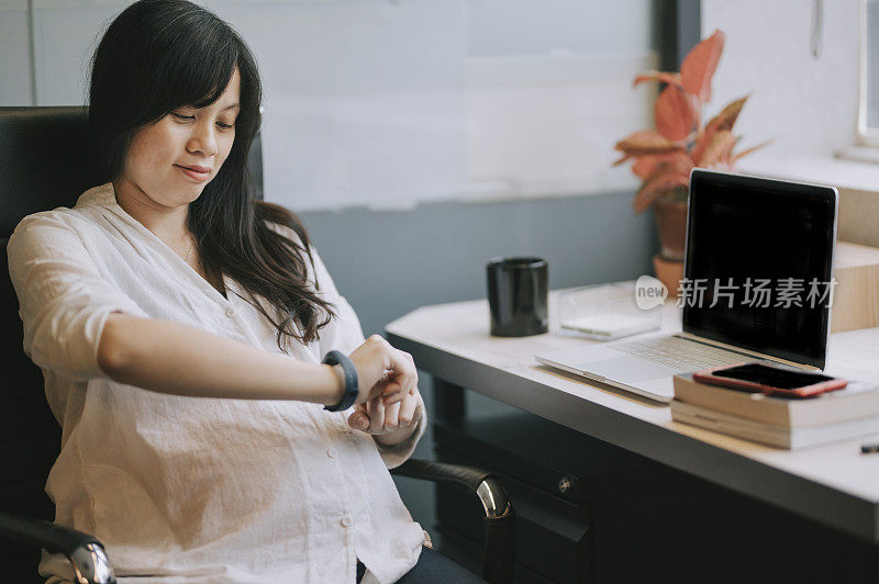 一名亚裔华裔孕妇在封锁期间在家工作，在家里的办公室查看她的智能手表