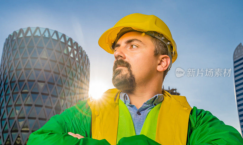 年轻的工程师戴着黄色安全帽和绿色防护服，双臂交叉，在现代化的商业建筑和建筑工地前看着摄像机。镜头光晕。低角度的观点