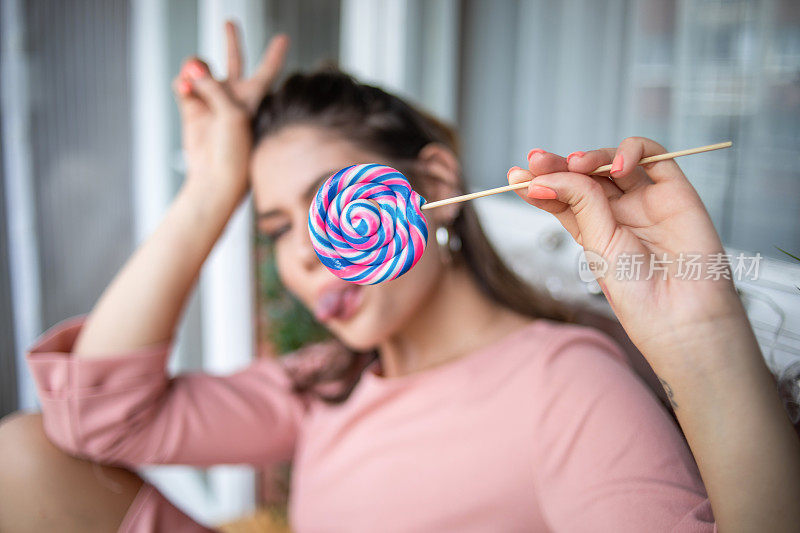 一位身穿粉红色连衣裙的美丽年轻女子坐在阳台上，手里拿着一根大棒棒糖。