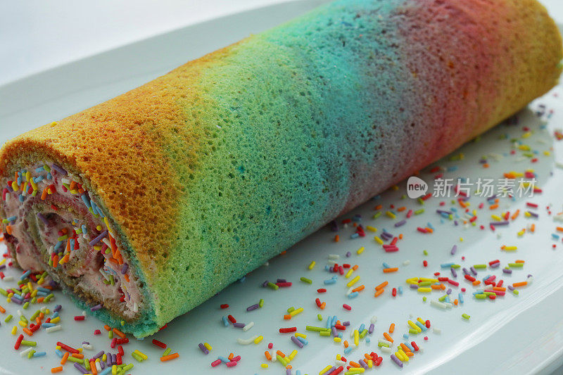 这是自制的彩色瑞士卷海绵蛋糕，里面有奶油和彩虹馅，白色的盘子里有彩虹色的条纹卷海绵蛋糕，周围有成千上万条糖丝，高架的视野