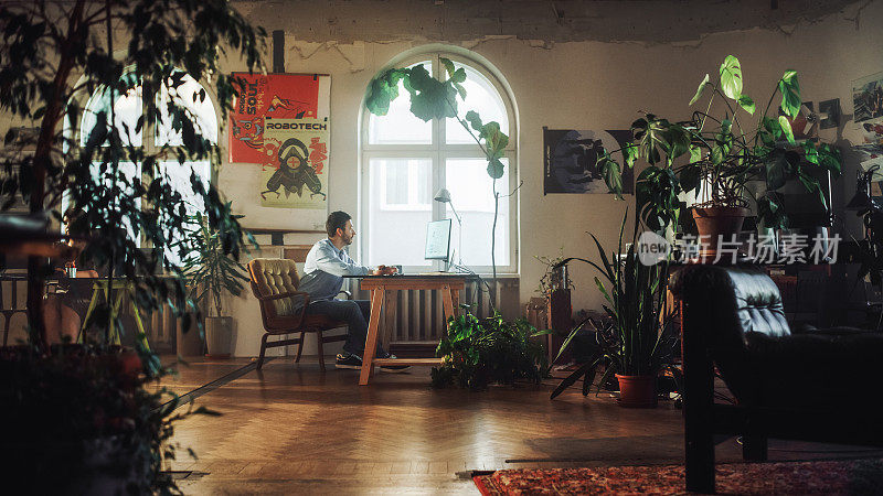 年轻英俊的男人在台式电脑上工作在创意机构在真实的Loft办公室。装修时尚的装饰与室内植物，艺术海报和大圆形窗户。