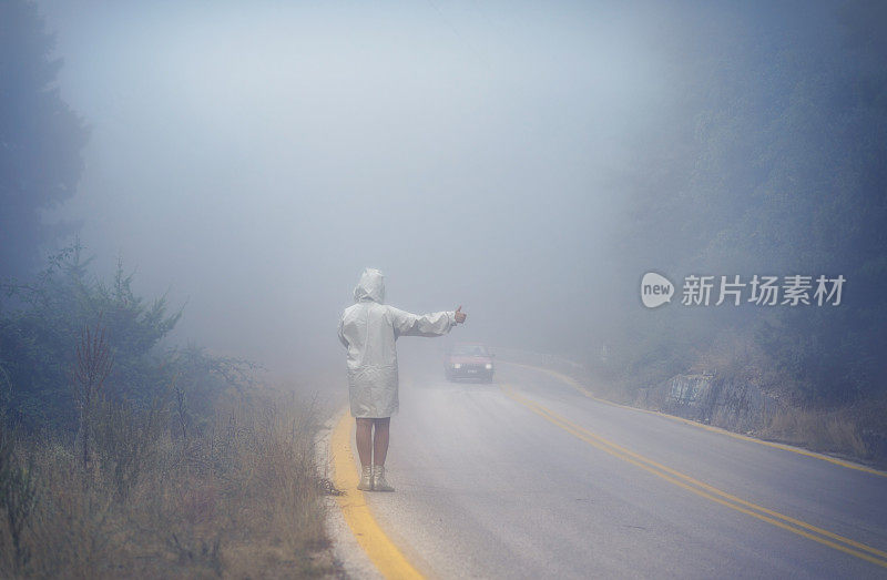 一名年轻女性穿着雨衣在雾中的道路上。穿着雨衣的女人在雨中搭便车旅行