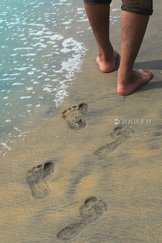 亚洲印度教印度男子光着脚走在印度的果阿沙滩上，金色的沙滩上有脚印，棕色的绿色牛仔裤卷起来，毛茸茸的腿被海浪打湿，溅起泡沫
