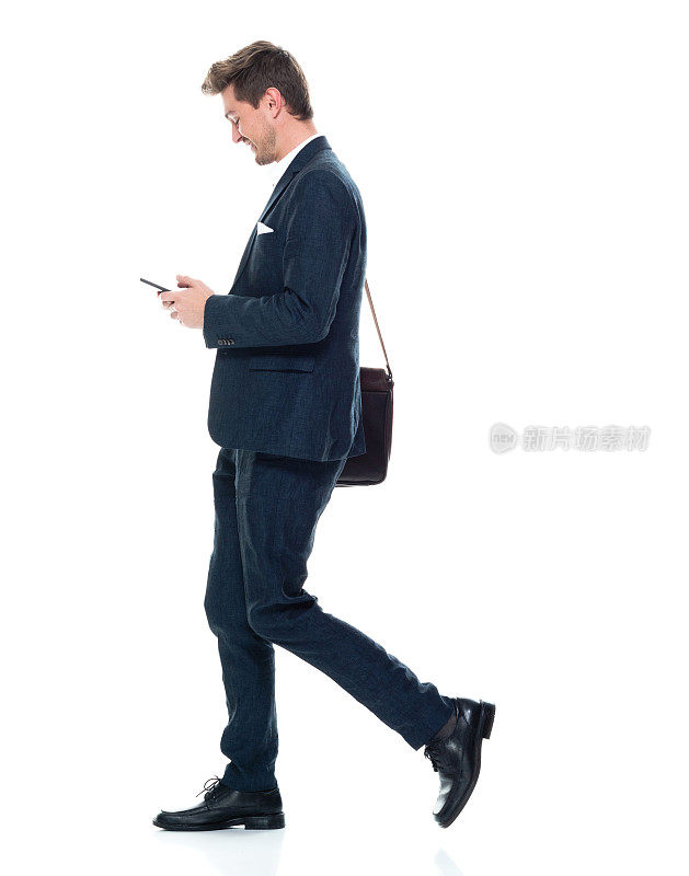 白人年轻男性经理走在白色背景前，戴着邮差包，拿着包，使用智能手机