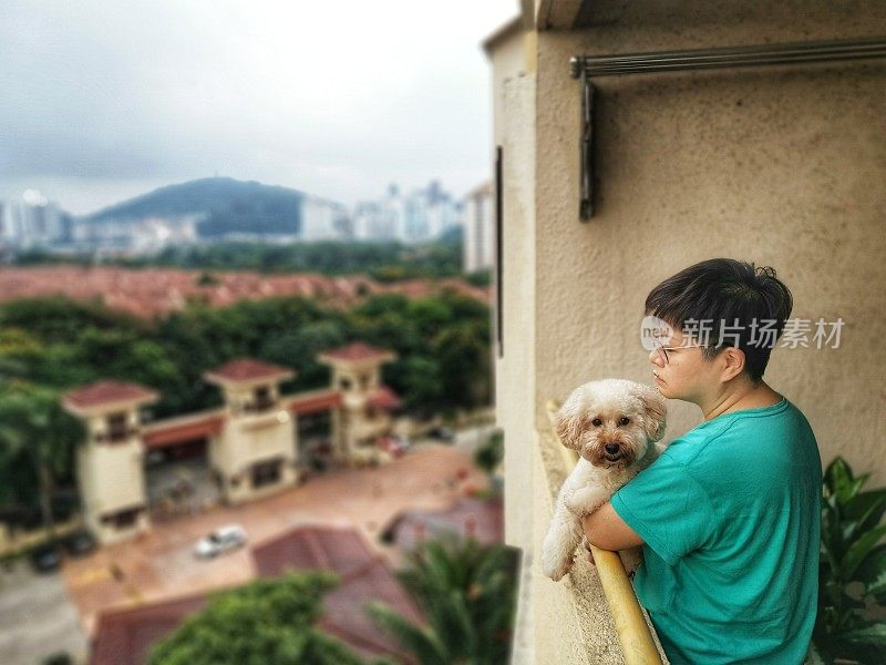 一名亚裔中国妇女抱着她的宠物玩具贵宾犬从阳台上望着她