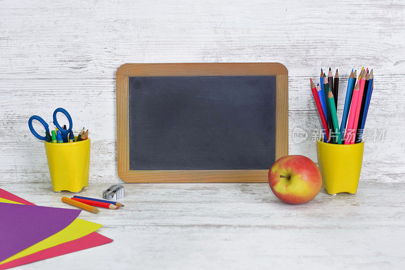 儿童书桌上有一块黑色的黑板，黄色的罐子里有几支彩色的铅笔和一个苹果