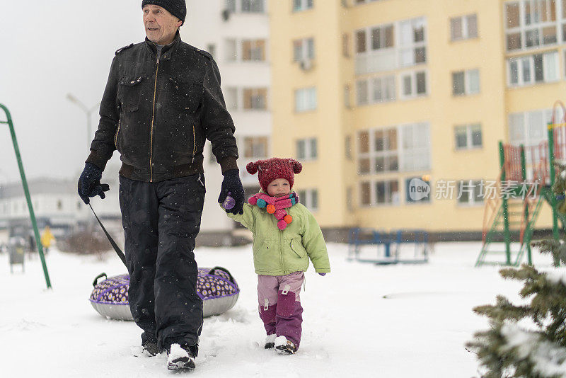 70岁的老人，爷爷和孙女扛着雪筒在雪中行走。