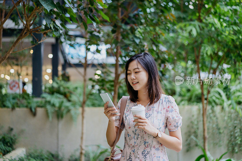 快乐的年轻亚洲女性使用智能手机和拿着纸咖啡杯