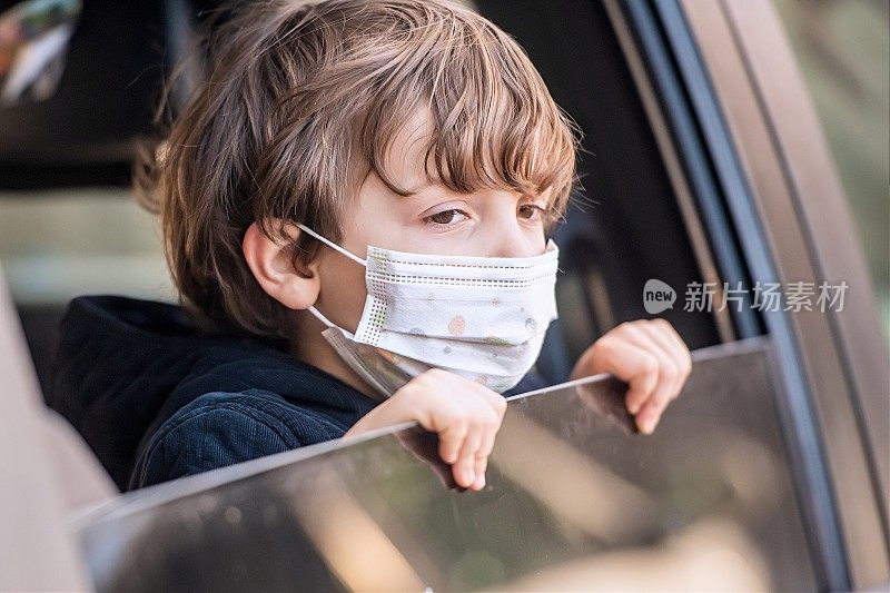 一个戴着防护面罩的小男孩在上学的路上，透过车窗看向别处