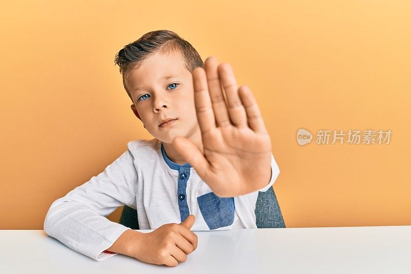 可爱的白人孩子穿着休闲衣服坐在桌子上做停止唱歌与手掌。警告表情与否定和严肃的手势在脸上。