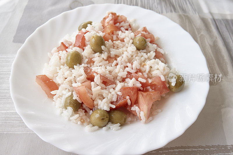 桌布上的白盘子上放着一盘凉拌米饭沙拉，上面有西红柿和橄榄。