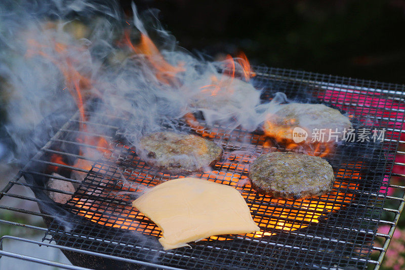 近景拍摄的是便携式的冒烟的火焰，花园里的木炭烧烤，在户外金属烤架上烤的汉堡，在汉堡肉饼上融化的加工过的奶酪片，聚焦前景