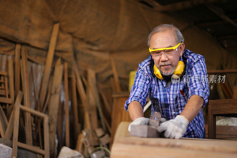 年老的亚洲木匠在他自己的车库风格的车间里用木板手工刨床爱好退休年龄