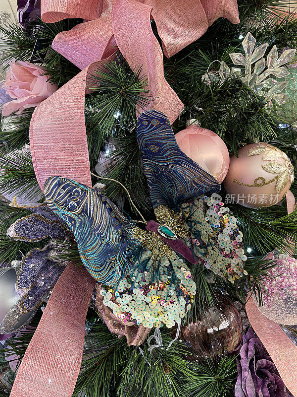 图像的亮片孔雀蝴蝶装饰挂在人造圣诞树的树枝上，粉彩丝带，蝴蝶结和小饰品，雪花，玫瑰花头，云杉针，模糊的白色仙女灯的背景
