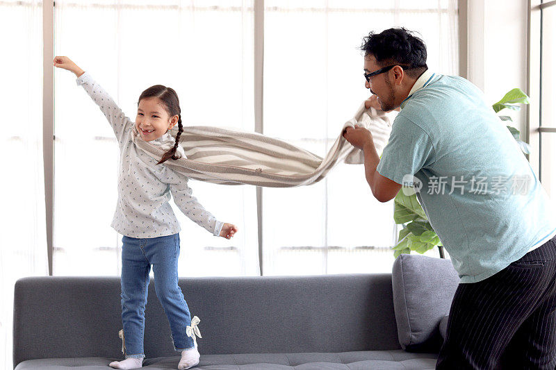 可爱的小女孩穿着大毛巾当超级英雄斗篷，爸爸抖着毛巾，假装她可以飞到客厅的沙发上，孩子和爸爸在家里一起扮演英雄，父亲节快乐。