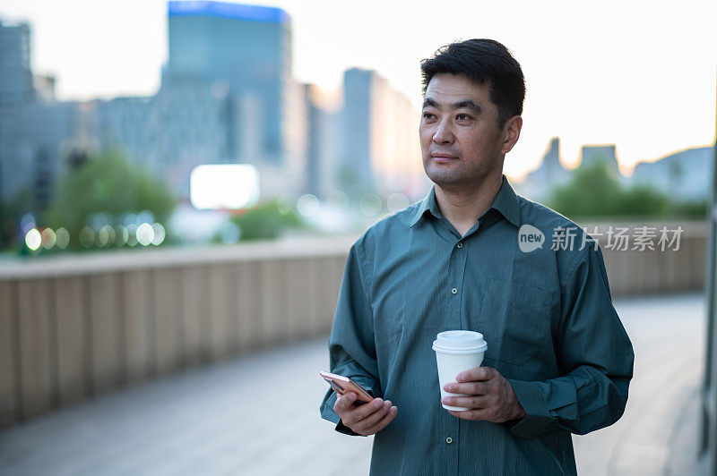 亚洲中年商人拿着咖啡杯，在城市街道上使用智能手机