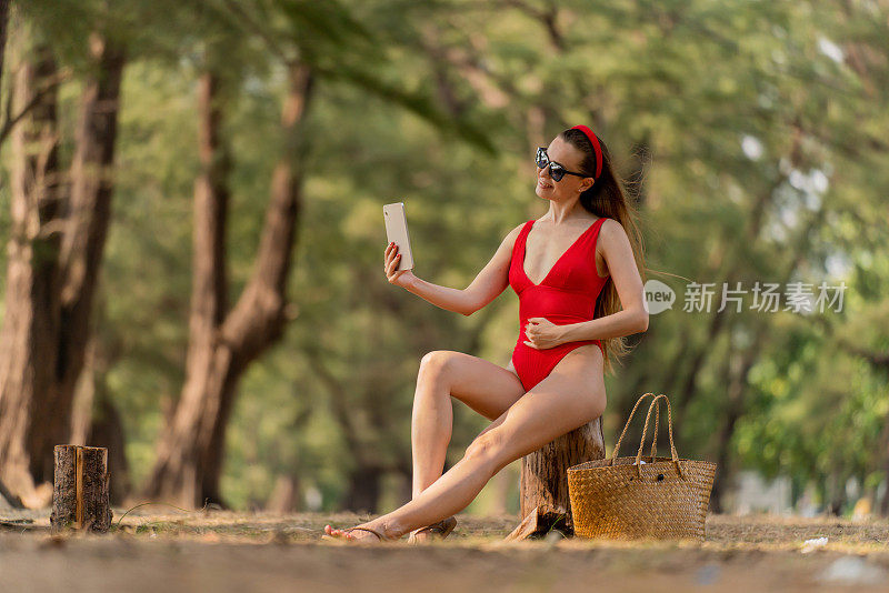 美丽的白人女性性感的女人在红色泳衣比基尼自拍与一个数字平板电脑，戴着太阳镜和一个红色的头带微笑凉爽和放松快乐的森林在海滩