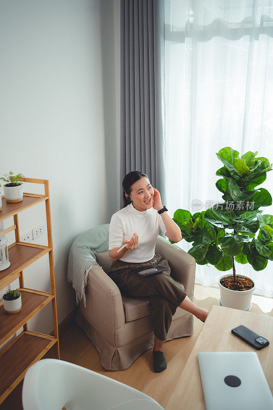 亚洲女性喜欢戴着耳机上网工作，在平板电脑上阅读数据，坐在家里的扶手椅上打电话。