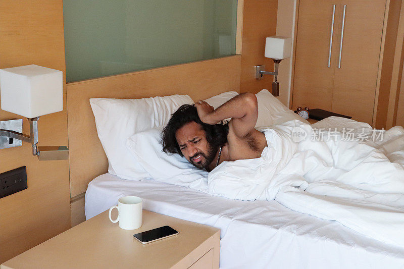 印度男子从羽绒被下望向酒店的双人床，看着床头柜上拿着咖啡杯的智能手机闹钟，挠头，困惑的表情，专注于前景