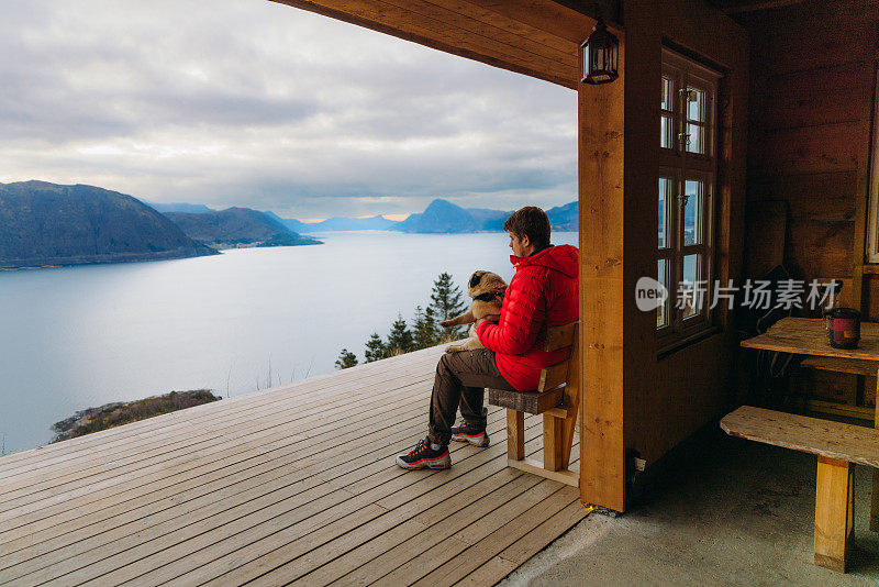 在挪威，一个快乐的男人带着狗在山上俯瞰风景优美的峡湾，沉思冬日