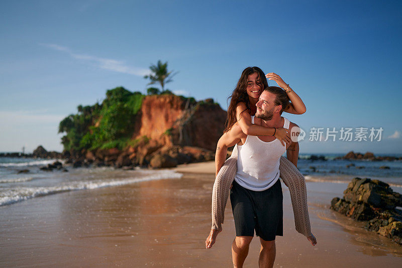 一对幸福的夫妇在热带海滩上度蜜月。男人快乐地背着女人。赤脚一副微笑，沿着岸边走。爱情，浪漫在海边绽放。新婚夫妇在日落时庆祝。