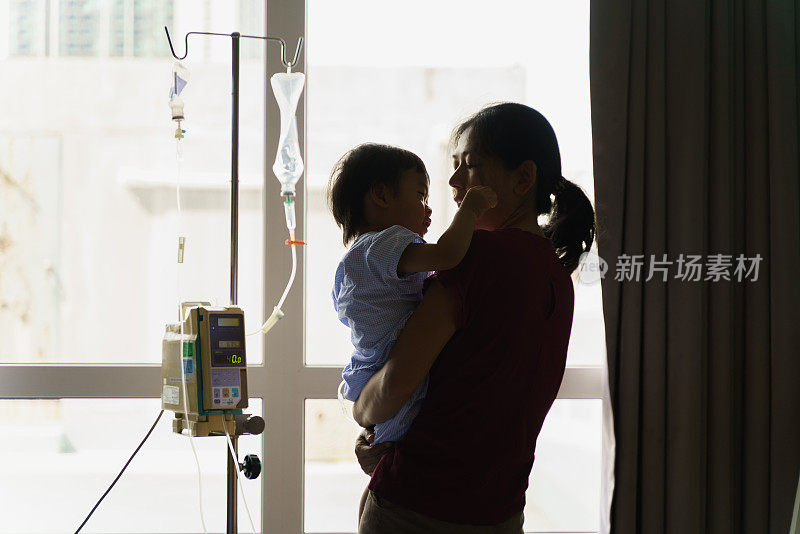 后视图剪影照片，亚洲母亲在医院康复时抱着并安慰她的儿子，手里拿着静脉点滴。可爱的亚洲孩子感染病毒住院了。婴儿保健医疗理念。