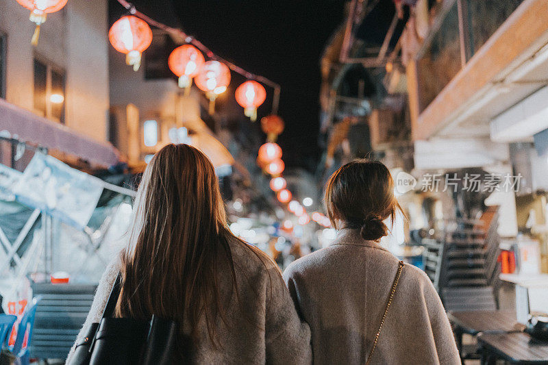 两名年轻的亚洲女子在香港长洲的春节庆祝活动中肩并肩走在一条似乎有商店的街道上。
