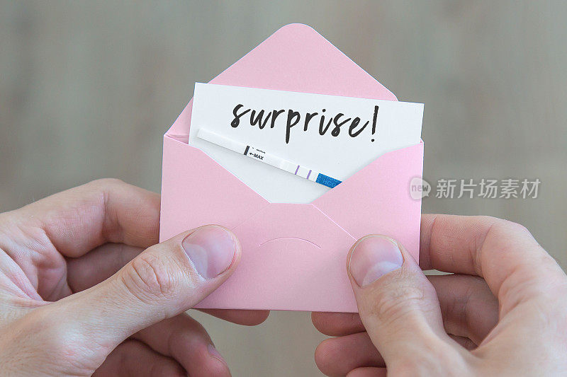 男性手里拿着一个粉红色的小信封，里面有一张明信片，上面写着惊喜和怀孕测试阳性，怀孕的概念