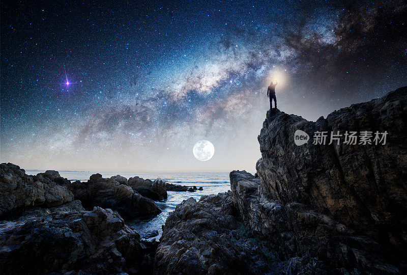 男子站在户外的岩石上，仰视，举起手凝视银河和月亮