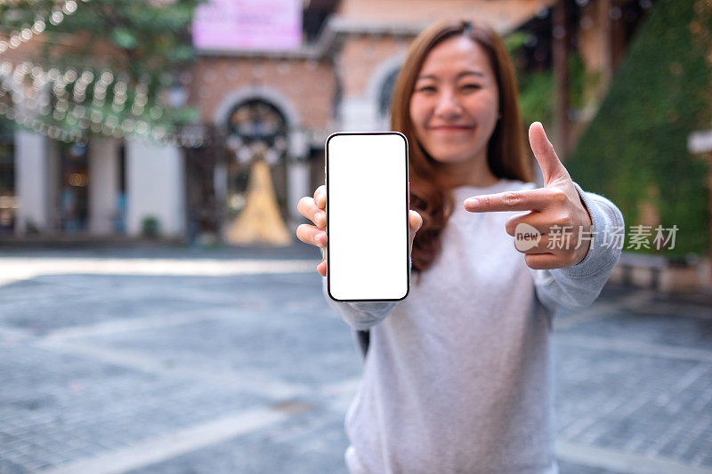 模拟图像的一个年轻的亚洲妇女持有和指着手指在户外的一个空白屏幕的手机