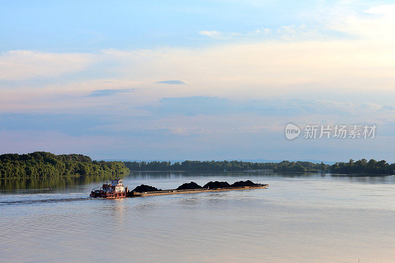 在多瑙河岸边的绿树丛中，一艘拖船拖着满载黑色煤炭的重型驳船