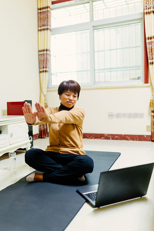 一位中年妇女在瑜伽垫上用笔记本电脑学习健身之道——科技与生活，健康的生活方式