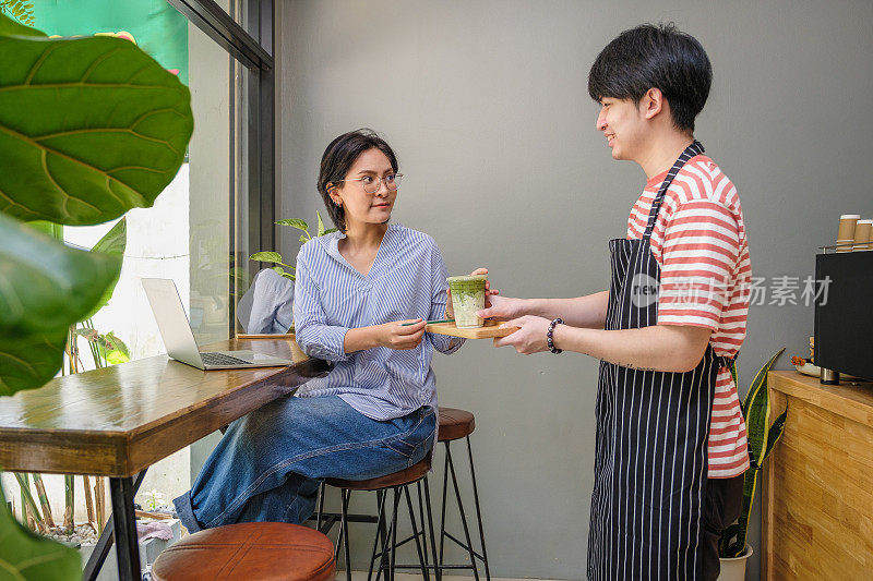 年轻的亚洲男子服务员在咖啡店的桌子上为他的顾客提供咖啡