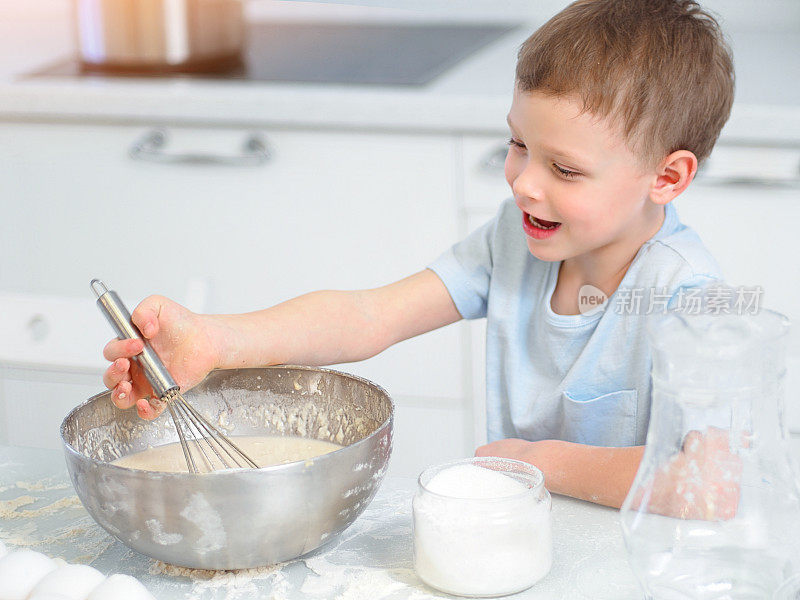 孩子在做饭。一个白人男孩坐在厨房的桌子旁，用手打蛋器在杯子里揉面。开朗积极的孩子做派，笑。年轻的厨师或厨师。孩子的肖像。