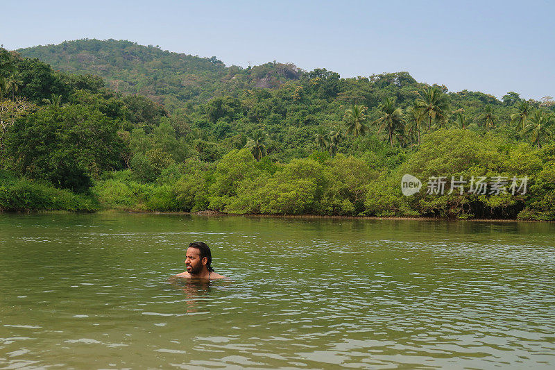 印度男子在泻湖平静的海水中游泳和戏水的图像，热带岛屿天堂，林地与棕榈树的景观，重点在前景