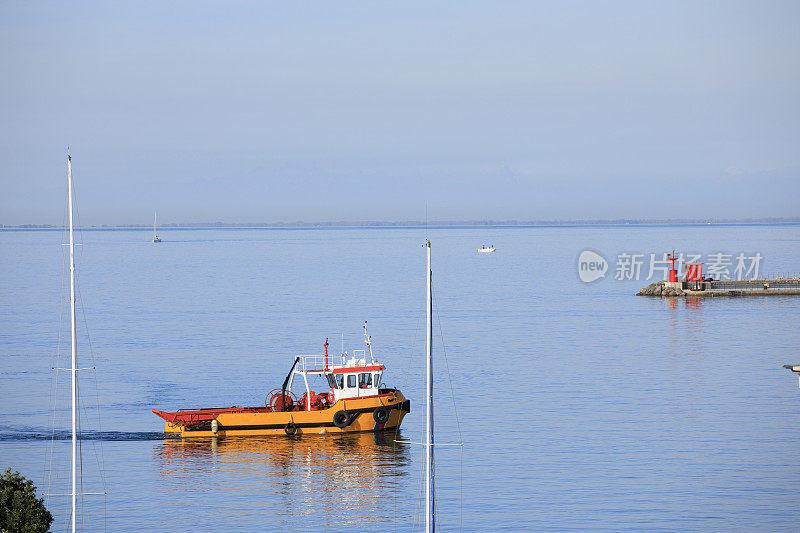 一艘黄色的小拖船，航行在平静的海面上，一个有红色灯塔的安全港口。亚得里亚海，的里雅斯特，意大利，欧洲。
