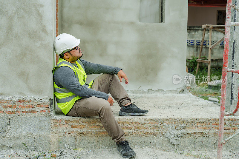 亚洲建筑师，建筑监理。戴上白色安全帽。靠着水泥墙坐着休息一下，疲惫的脸，满头大汗。穿着反光安全背心的男子。