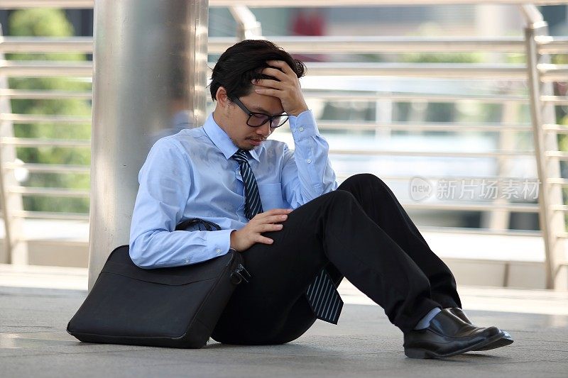 一幅沮丧沮丧的亚洲年轻人的肖像，他坐在人行道上的办公室地板上，对工作感到疲惫