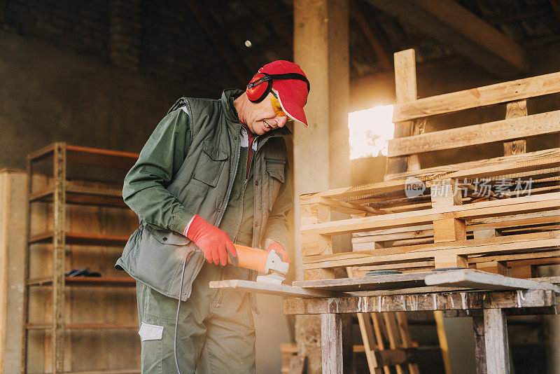 图为成熟的木匠穿着防护服，用电动研磨机打磨木材。在阳光明媚的日子里，在他的车库里享受他的工作。