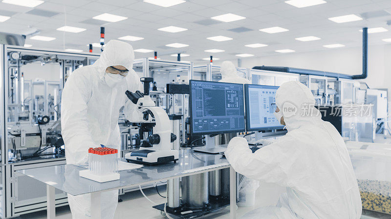 一队研究科学家在无菌服工作与计算机，在显微镜下看和现代工业机械在实验室。产品制造工艺:制药，半导体，生物技术。