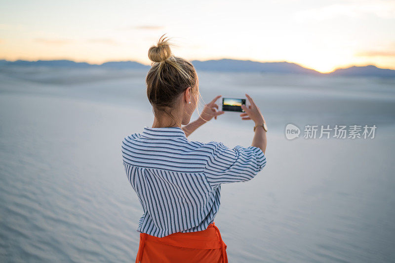 年轻女子使用智能手机自拍站在美丽的白色沙丘日落，时髦的女孩拍摄日落享受旅游度假的风景