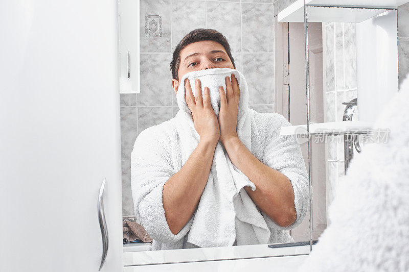 一个穿着白色浴衣的男人在浴室的镜子里用白毛巾擦脸。