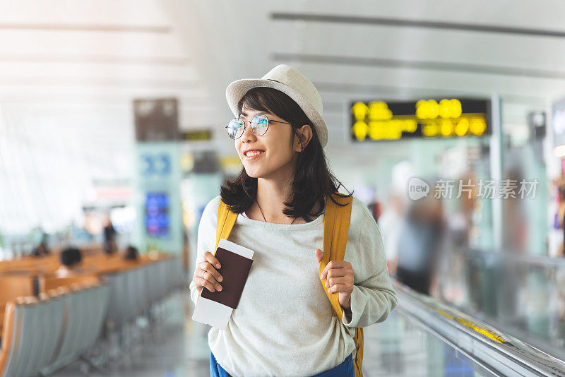 亚洲女子戴着眼镜、帽子戴着黄色背包正拿着飞机票、护照在机场大厅里。