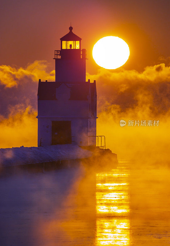 在寒冷的冬季空气中，灯塔在冒着蒸汽的湖面上日出。