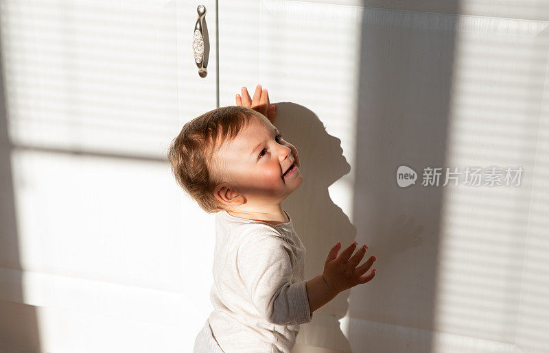 快乐的宝宝迈出了第一步，双手平衡在白色的厨房背景和早晨的太阳落在他身上