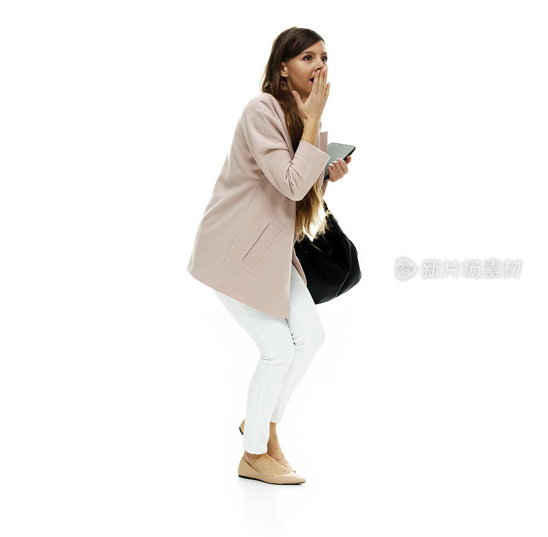 白种人年轻女性穿着外套，手持钱包，在白色背景前使用数码平板电脑