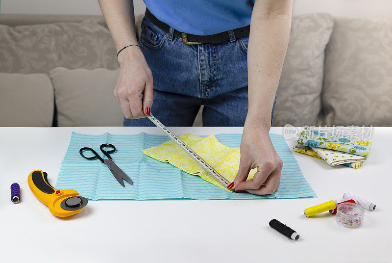 穿蓝色t恤的女人量布料。桌子上有缝被子的工具。拼布刀、剪刀、衬里剪垫、自锁、线、厘米胶带。缝纫用品