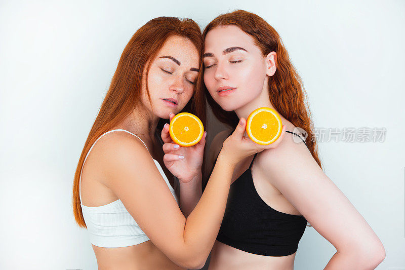 两个红头发的年轻女子都拿着一半的橙色在他们的手面对对方站在孤立的白色背景，美丽肖像的概念